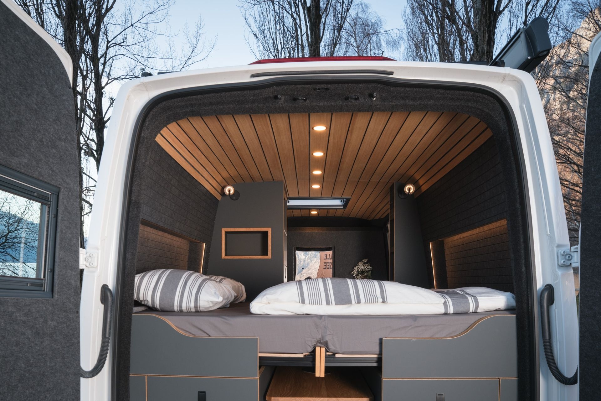Innenansicht von Campervan mit Hecktüren geöffnet. Doppelbett mit weisser Bettdecke. leichte beleuchtung an Wand und Decke