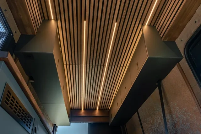 Moderne Camper Van Decke von Cocoon Vans mit Holzverkleidung aus amerikanischem Nussbau und integrierten dimmbaren LED-Lichtleisten, mit eleganten grauen Hängeschränken auf beiden Seiten.