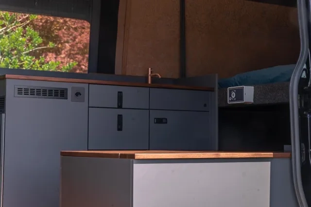 Blick auf die Küche im Camper Van. Das Hubbett ist herunter gelassen. Die Sonne scheint auf die Oberflächen aus amerikanischen Nussbau.